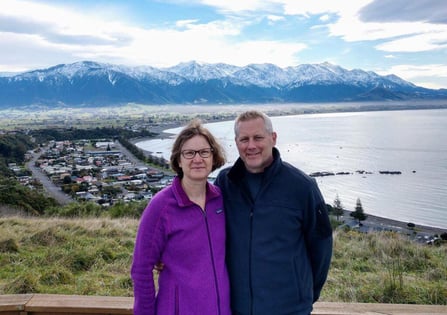 A man and a woman standing on an overlook with a small town, an ocean, and the mountains behind them.