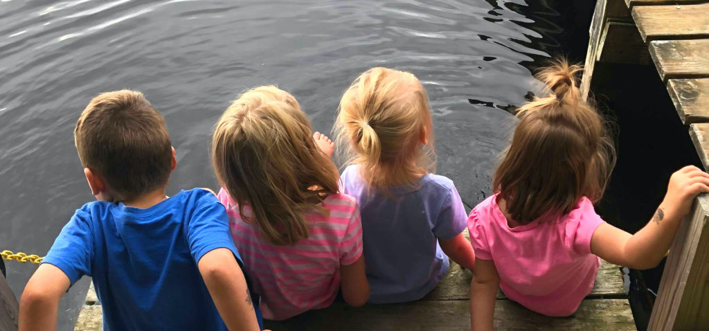 Four children sitting on a dock by a lake.