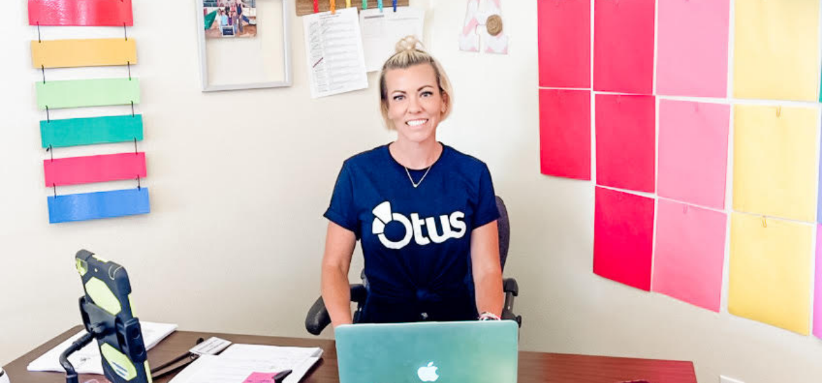 A woman wearing an Otus shirt sits at her desk in a classroom. The walls and her desk are brightly decorated and she sits in front of a Mac.